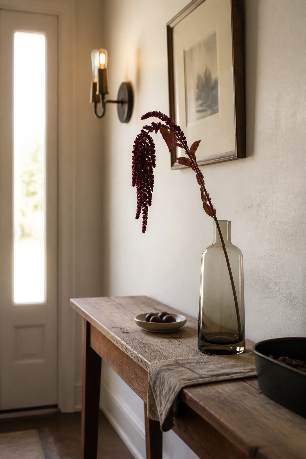 Wooden console table with smoky glass vase of drooping burgundy flowers near door.