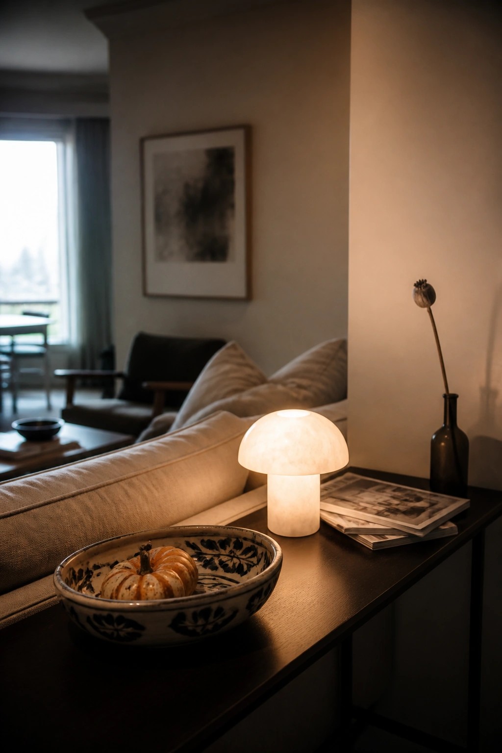A glowing mushroom lamp rests on a dark wooden console next to a patterned bowl holding a small pumpkin, with magazines and a dark vase nearby.