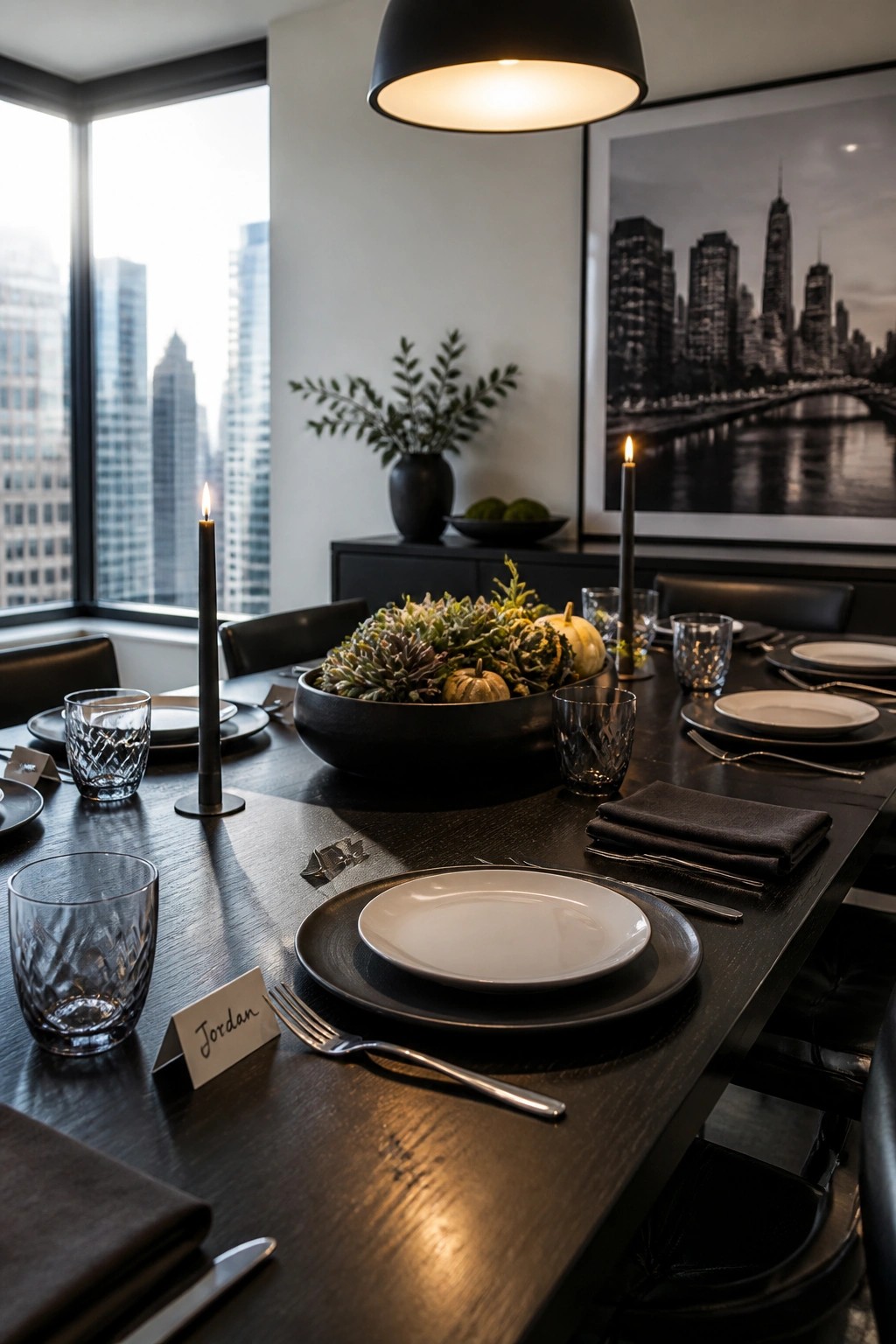 A modern dining table set with white plates on black chargers, black candles, a dark bowl centerpiece holding succulents and small pumpkins, and name cards beside the place settings.