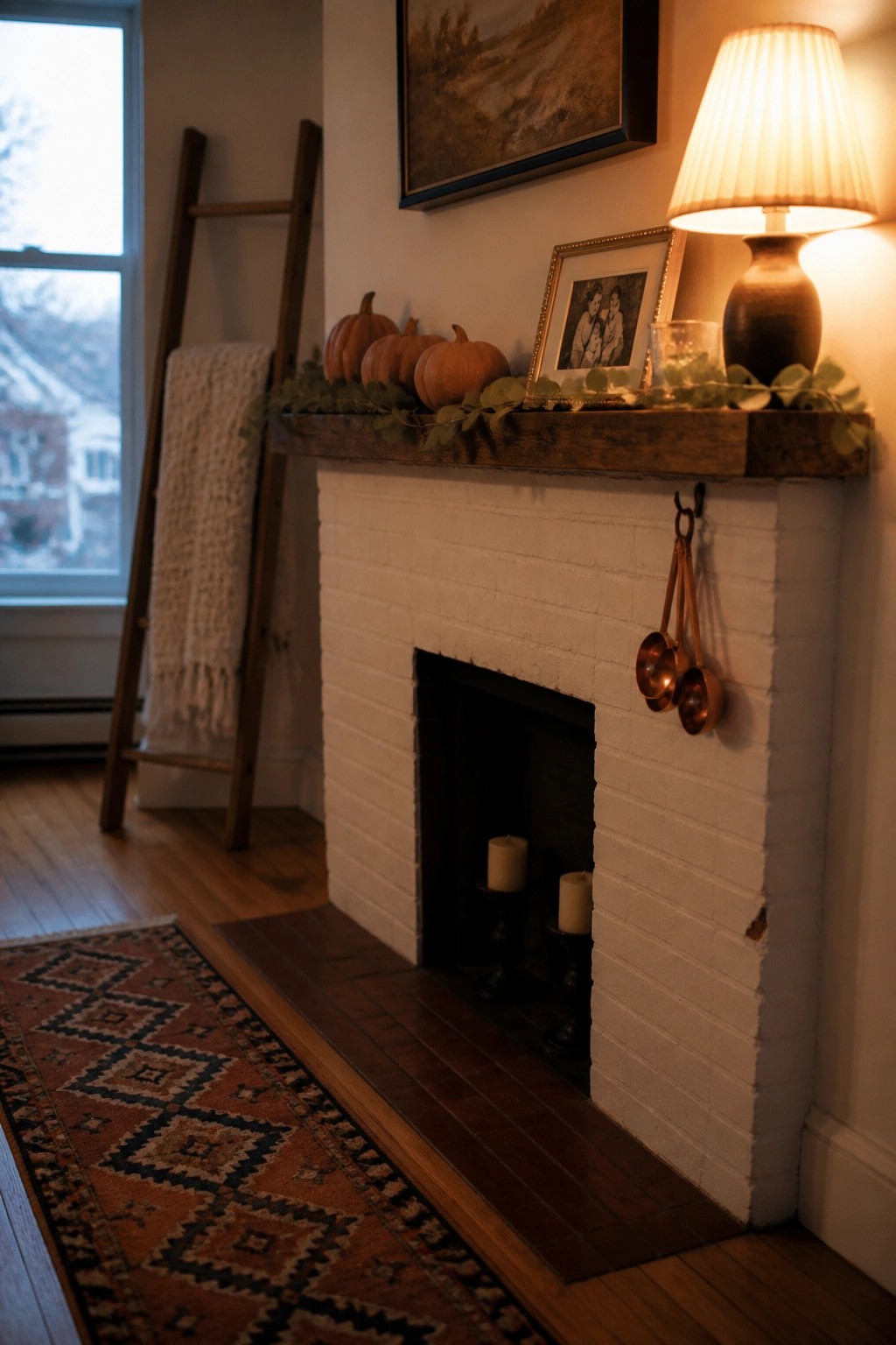 A white brick fireplace with a rustic wood mantel decorated with orange pumpkins, green foliage, framed photos, a table lamp, and hanging copper measuring cups.