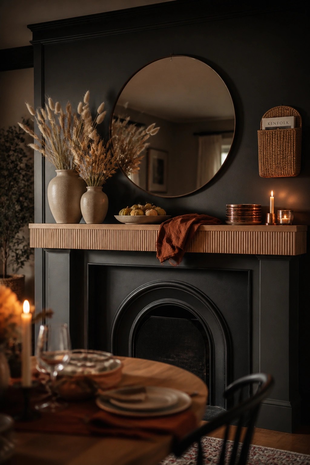 A black fireplace mantel topped with two ceramic vases of dried grasses, a bowl of small pumpkins, a folded rust cloth, stacked copper plates, and lit candles, with a round mirror centered above.