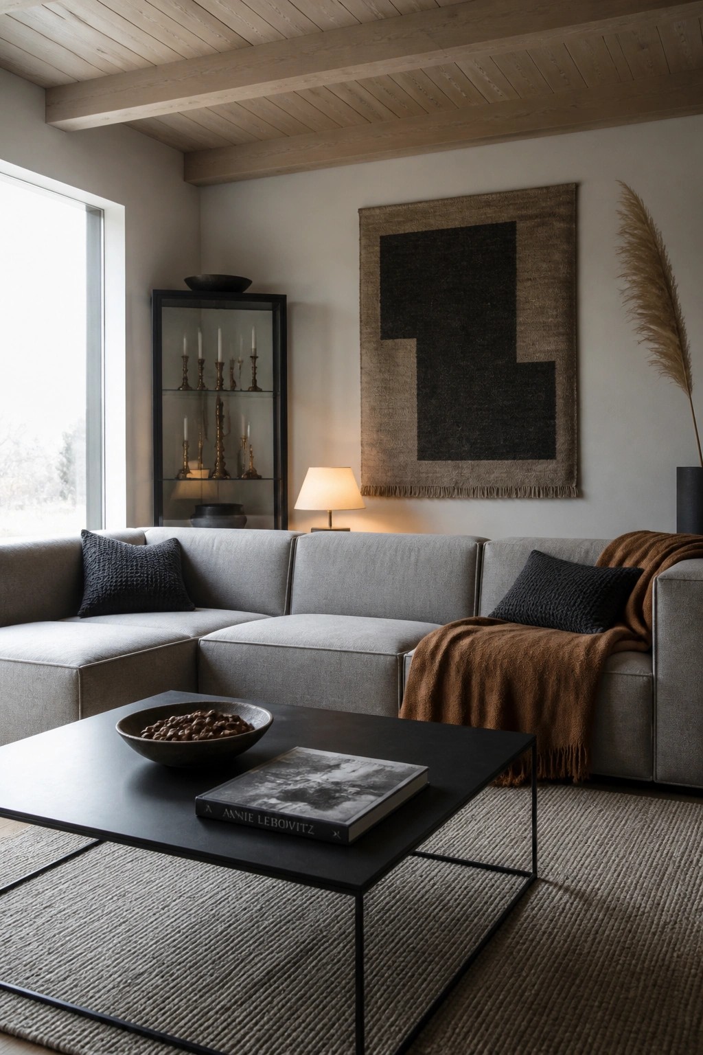 A modern living room with a gray sectional sofa, dark coffee table holding a bowl and book, wall tapestry, and display cabinet.
