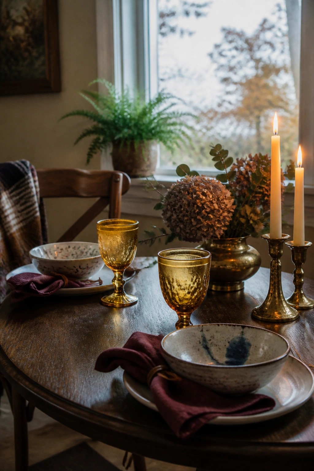 A dark wooden dining table set with amber glass goblets, burgundy napkins, speckled ceramic bowls, brass candle holders with lit tapers, and dried hydrangeas in a vase.