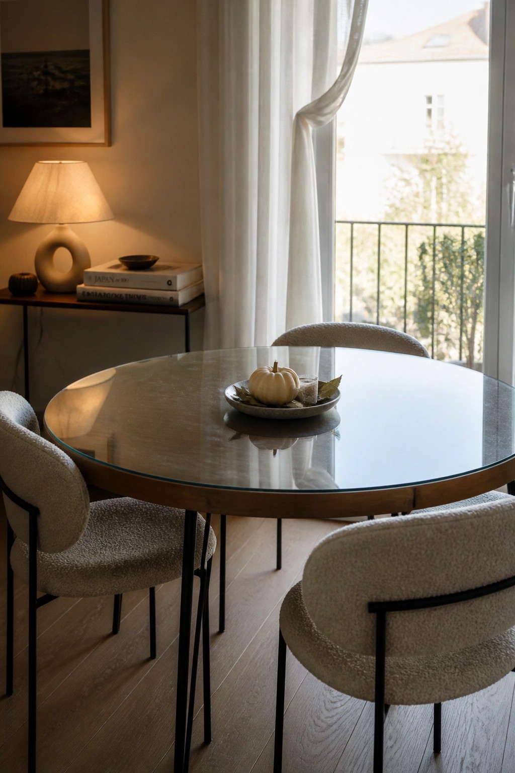A round glass dining table holds a small white pumpkin and leaves on a plate as its only centerpiece, with four light upholstered chairs around it in a room with wood floors and a window.