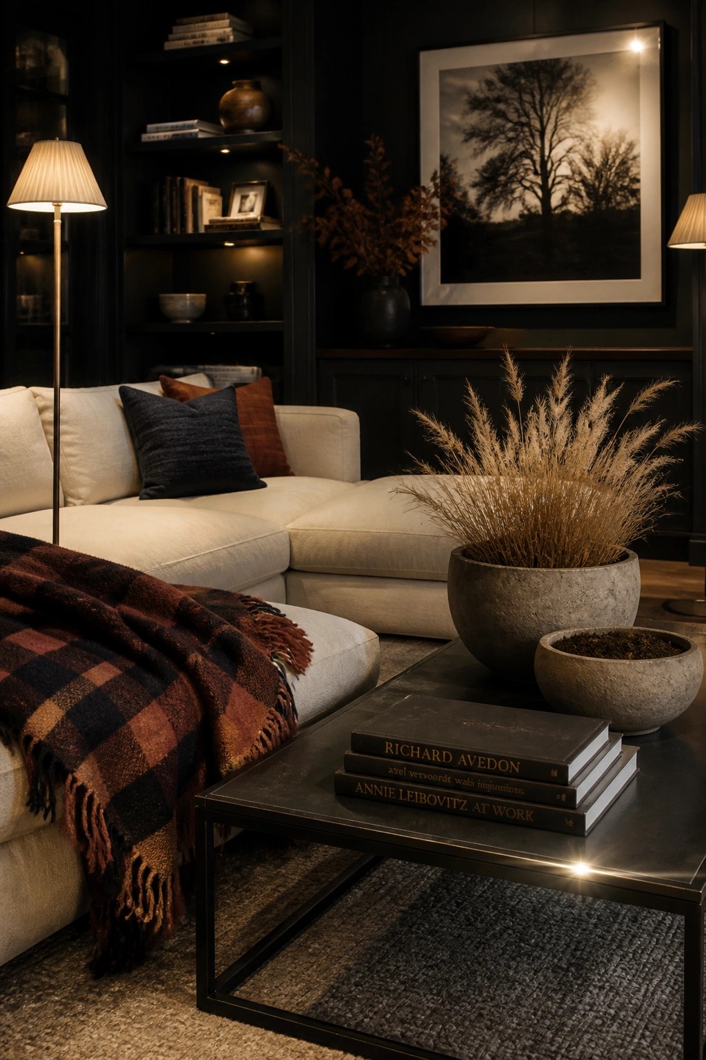 A living room scene showing a light sectional sofa with a plaid throw, pillows, a dark coffee table with stacked books, and large stone pots holding dried grasses.