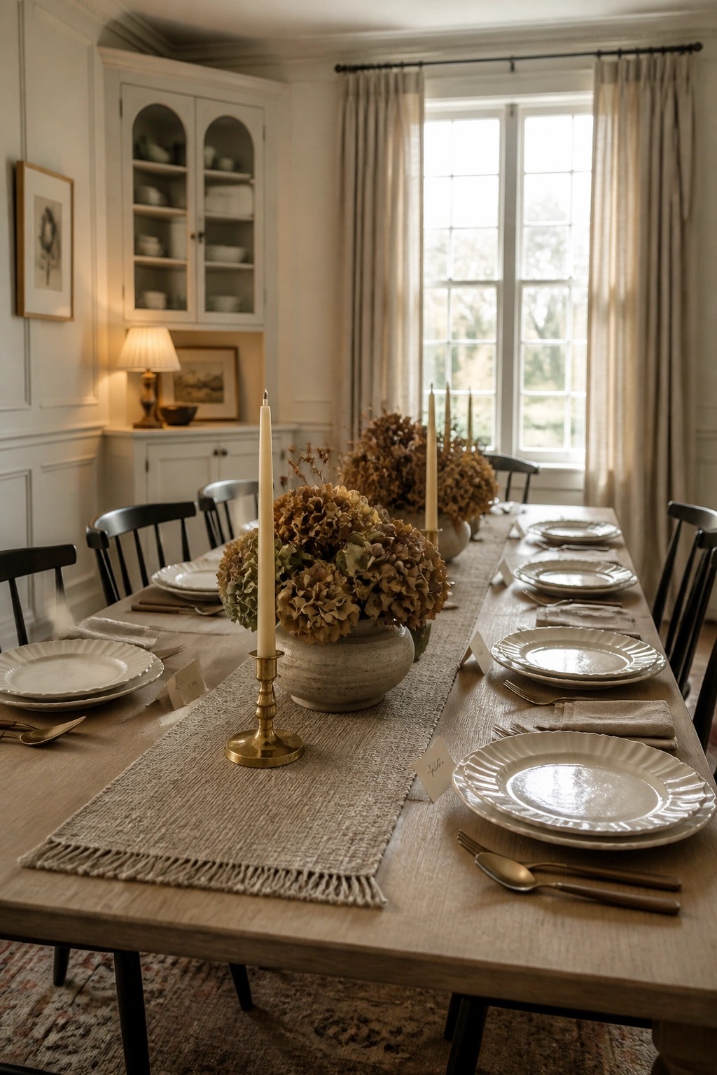 A dining table set with white plates, beige napkins, dried hydrangeas in stoneware vases, brass candle holders, and a long fringed linen runner.