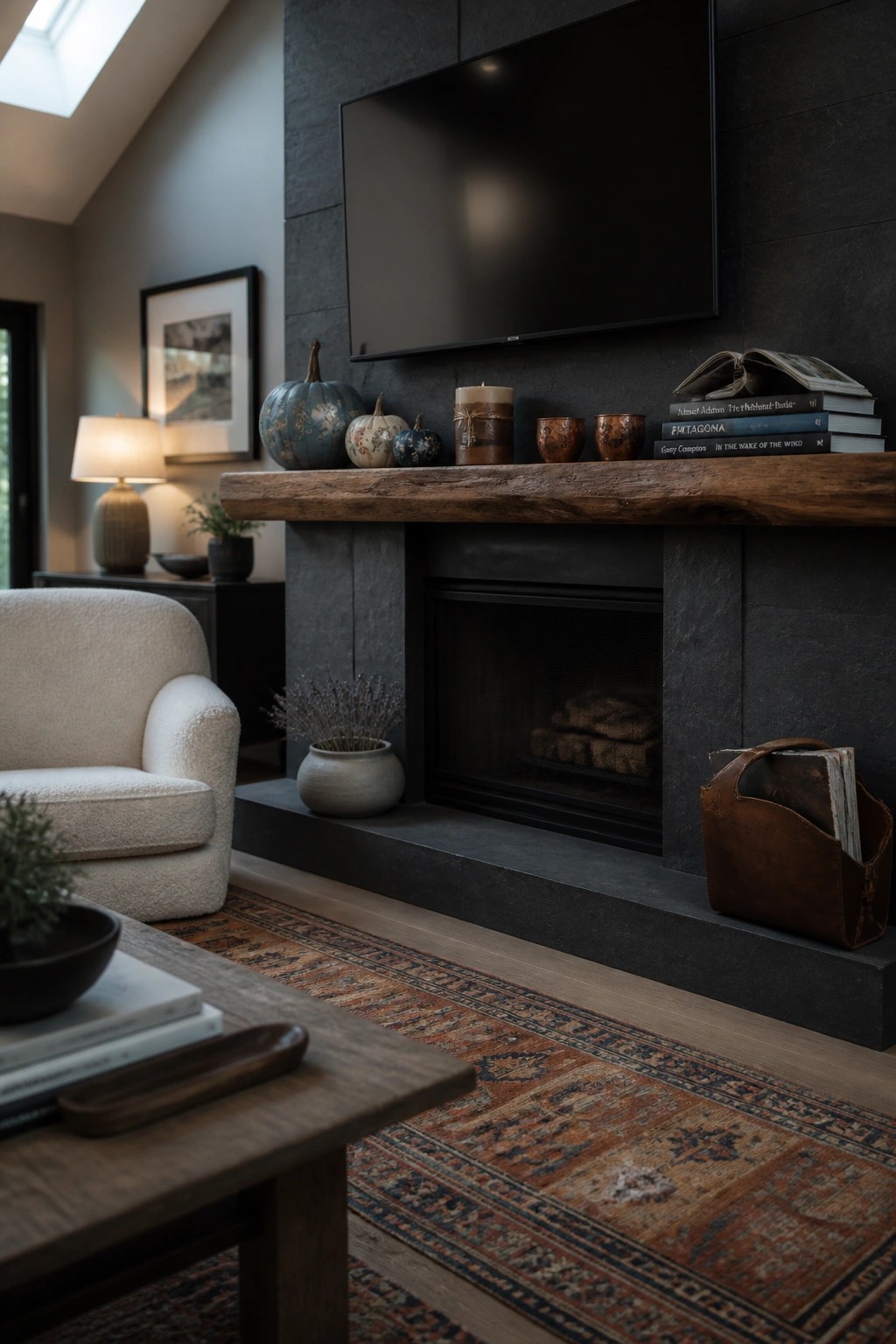 A dark stone fireplace with a wooden mantel holding small pumpkins, candles, books, and a potted plant, with a television mounted above.