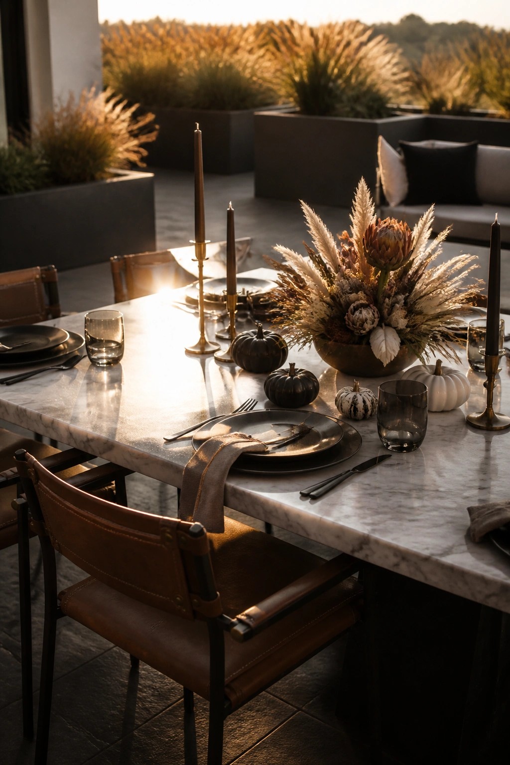 A marble outdoor dining table set with dark plates, glasses, candles, small pumpkins, and a large centerpiece of dried grasses and flowers.