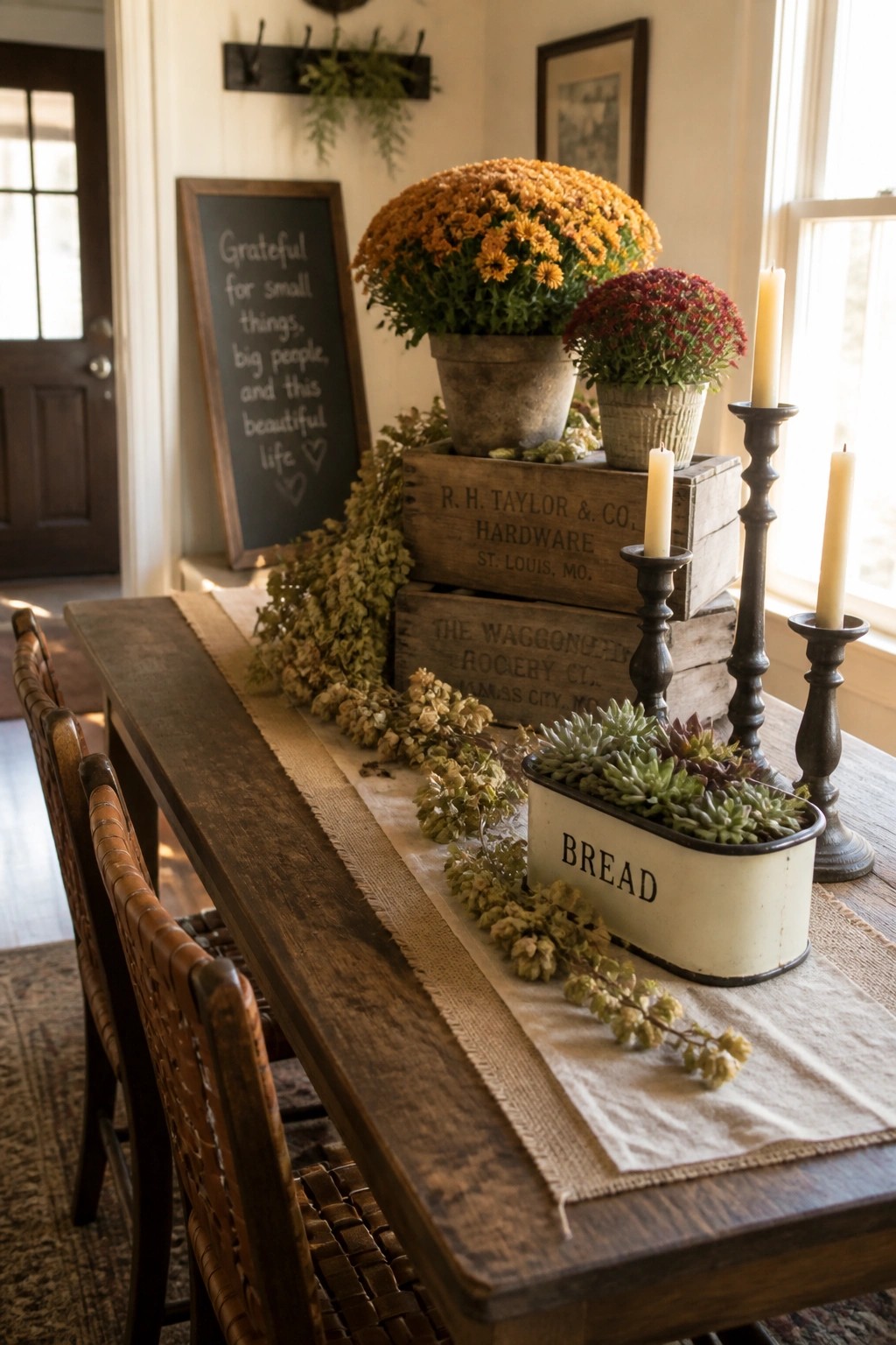 A wooden dining table with a fall centerpiece of stacked wooden crates holding potted orange and red chrysanthemums, black candlesticks with tall candles, and a white bread box filled with succulents, all arranged on a burlap runner.
