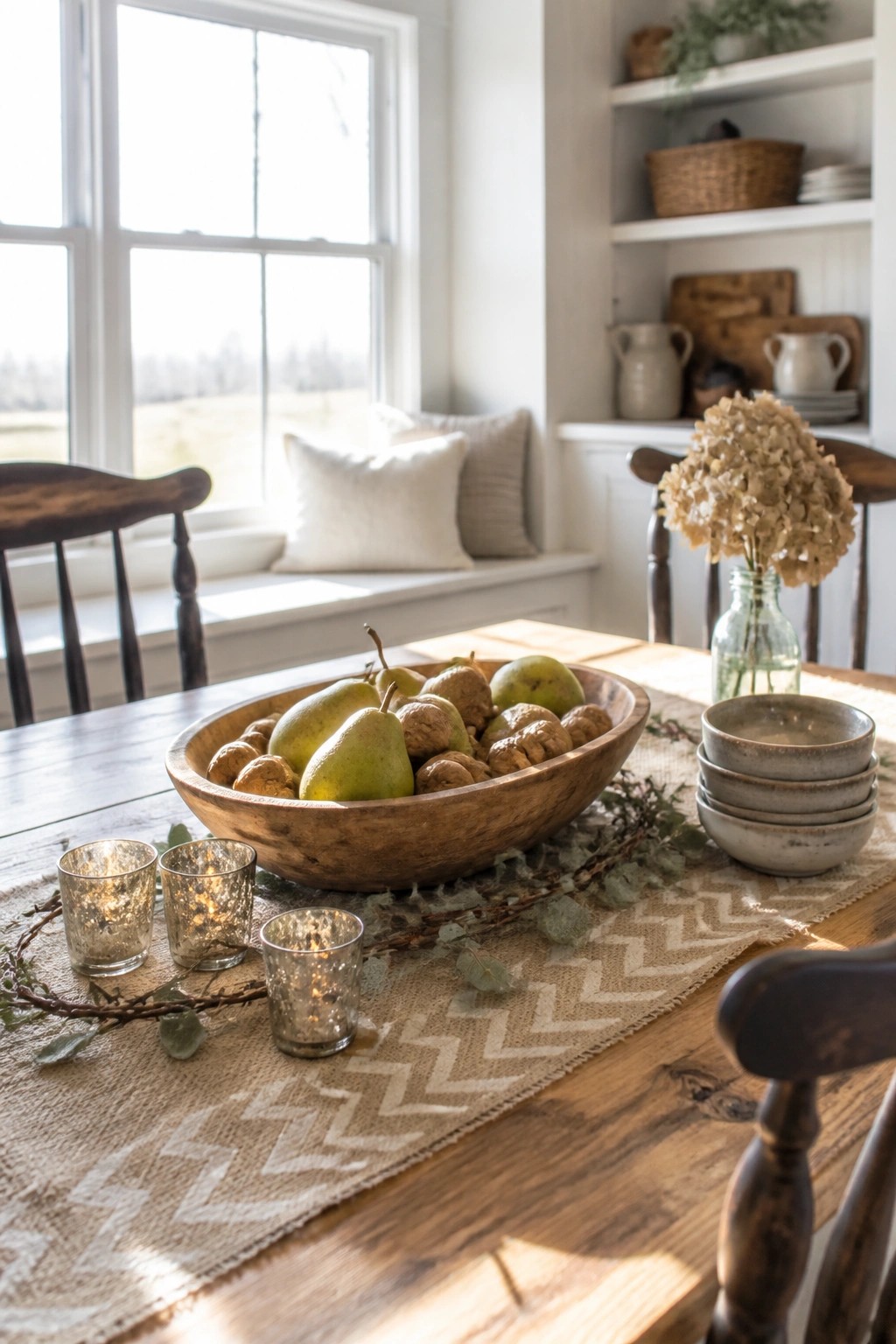 A wooden bowl of pears and walnuts centers a rustic dining table with a burlap runner, votive candles, and dried flowers nearby.