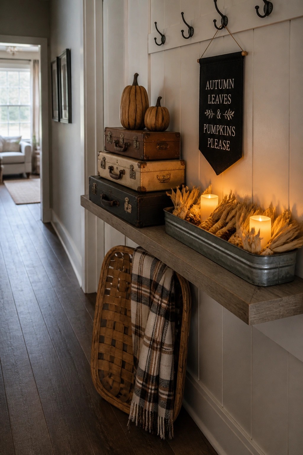 A narrow wooden shelf holds three stacked vintage suitcases with two small pumpkins on top, next to a metal tray containing lit candles and dried corn husks, with a black fabric banner hanging on the wall above.
