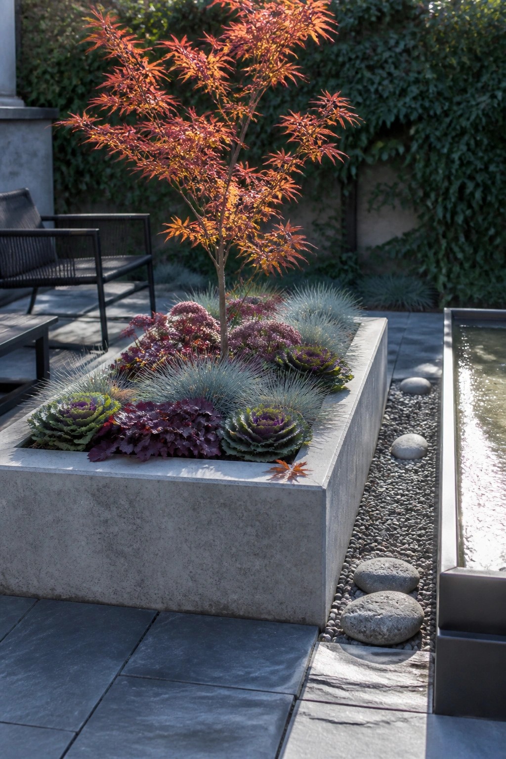 A concrete raised planter filled with a small red-leafed tree, purple cabbages, and blue-green grasses sits beside a gravel path and water feature.