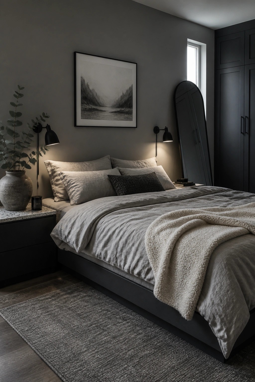 A bedroom with dark gray walls, a black bed frame, layered light gray and beige bedding with a textured throw, two black wall sconces, and a large framed landscape print above the bed.