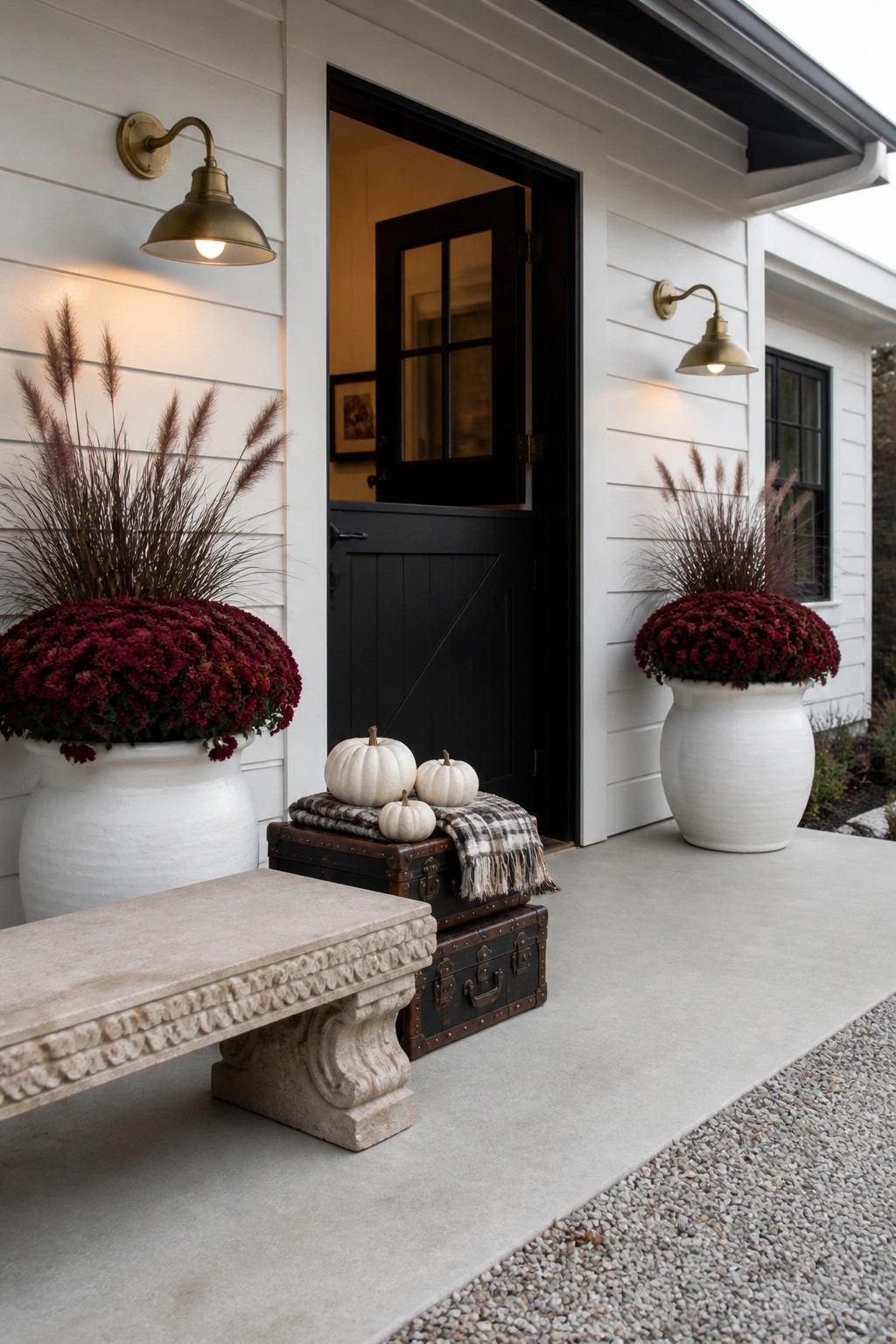 A white house exterior with black front door flanked by two large white planters filled with deep red chrysanthemums, a stone bench on the left, and stacked trunks holding small white pumpkins in front of the door.