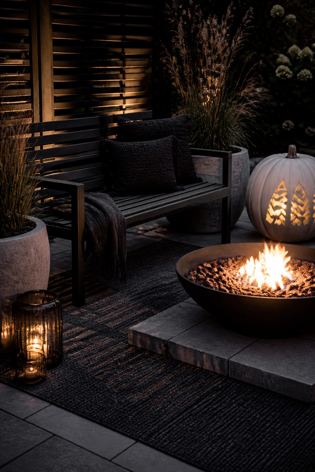 An outdoor patio at night with a lit fire pit in a dark bowl, a wooden bench with dark cushions, glowing glass lanterns, and a carved white pumpkin.