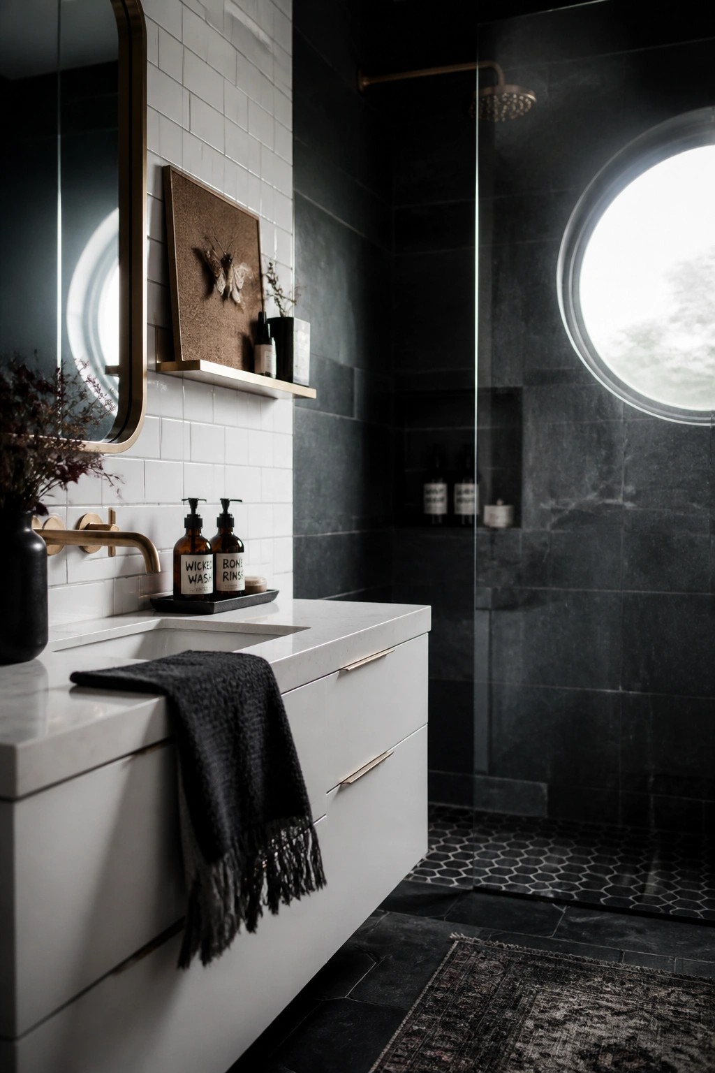 A modern bathroom with black tiled walls and floor, a white floating vanity with brass fixtures, a round mirror, and a glass shower with a circular window.