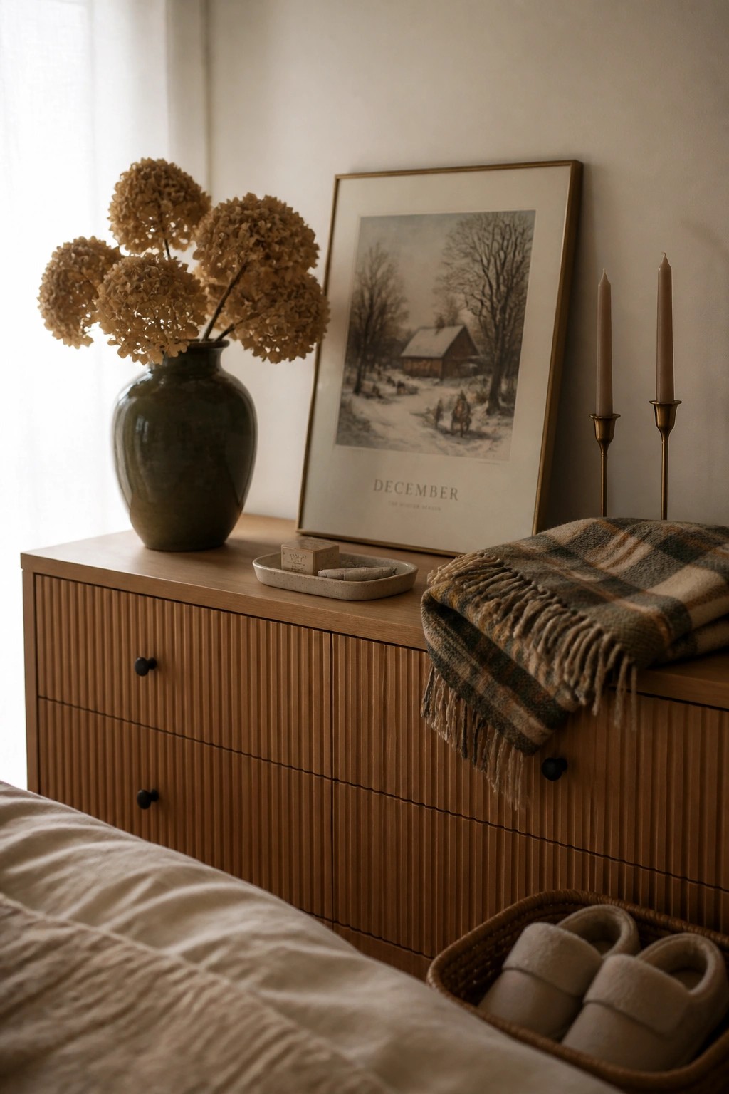 A light wood dresser with a green vase of dried hydrangeas, a framed winter print, two taper candles, and a plaid throw draped over the right side, plus a basket of slippers on the floor below.