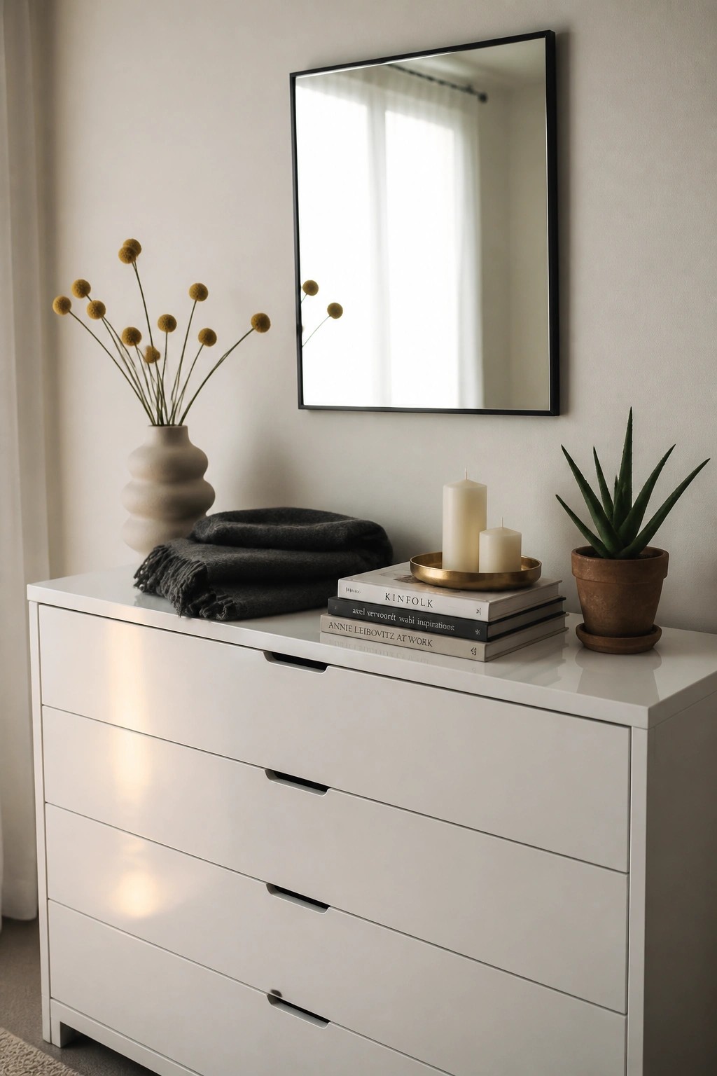 A white dresser with a mirror above it holds stacked books, two candles on a tray, a vase of dried flowers, folded blankets, and a potted plant.
