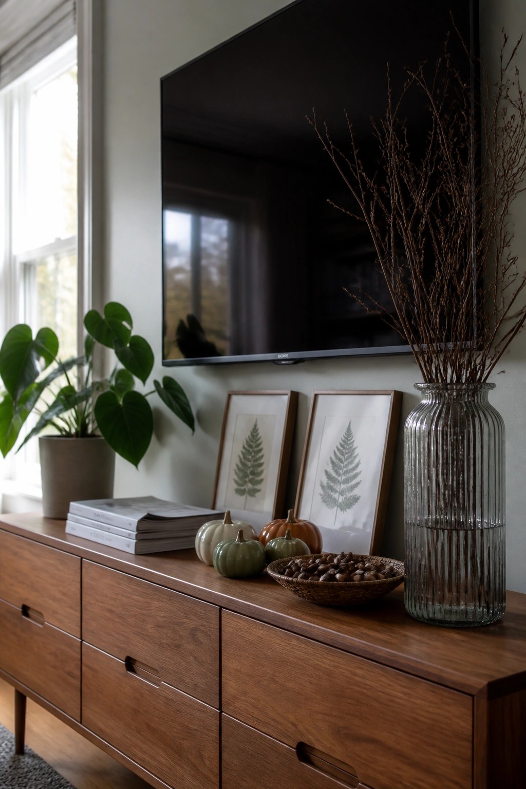 Wooden console under mounted TV with pumpkins, fern prints, books, and branch-filled vase
