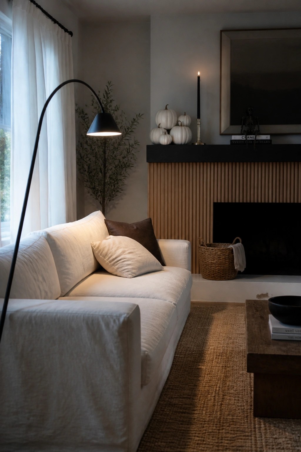 A minimalist living room with a white sofa, wooden coffee table, and fireplace mantel holding white pumpkins and a lit black candle.