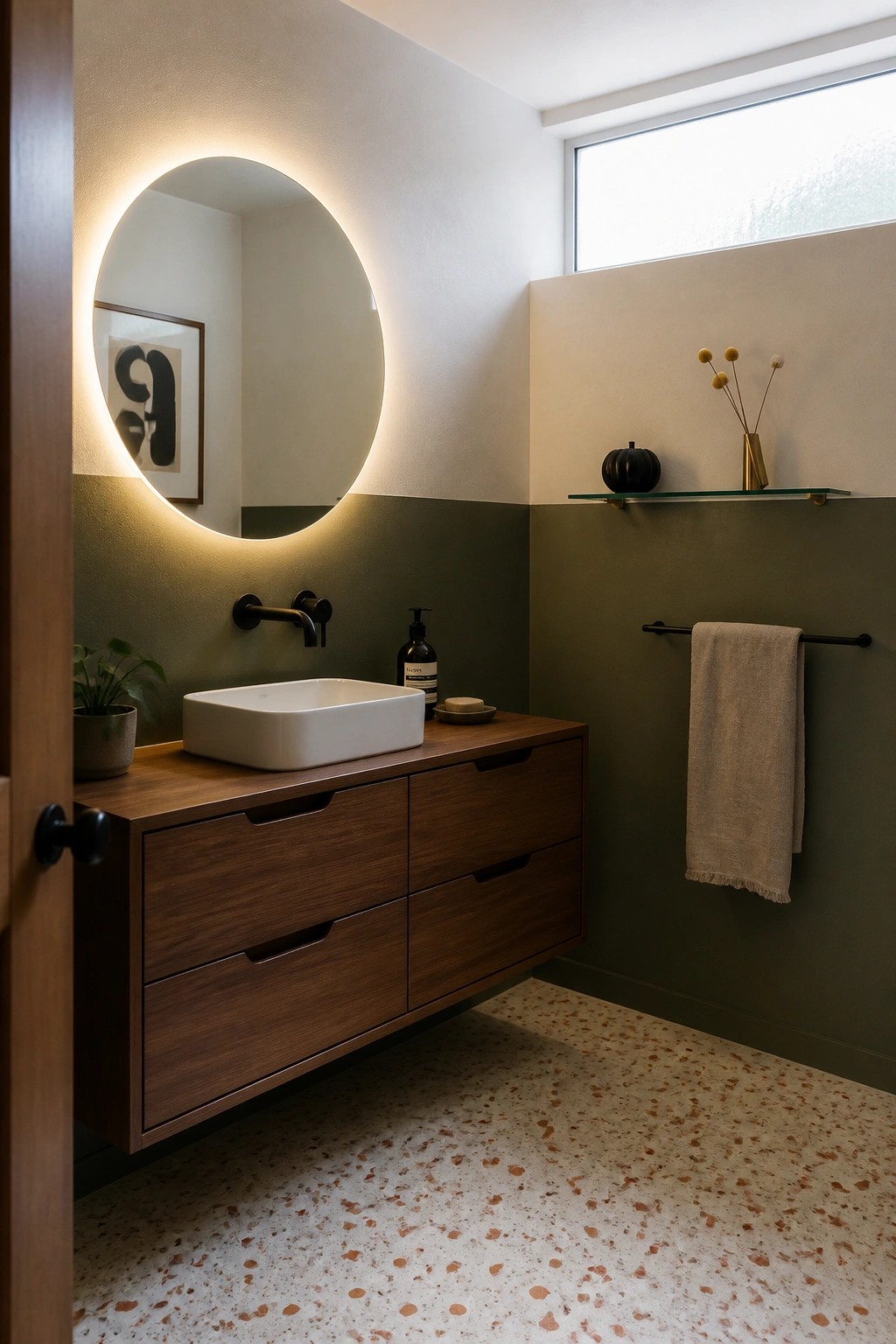 A bathroom interior showing a white floating sink on a wood vanity, round backlit mirror, green two-tone walls, and a glass shelf with a small black pumpkin and dried stems.