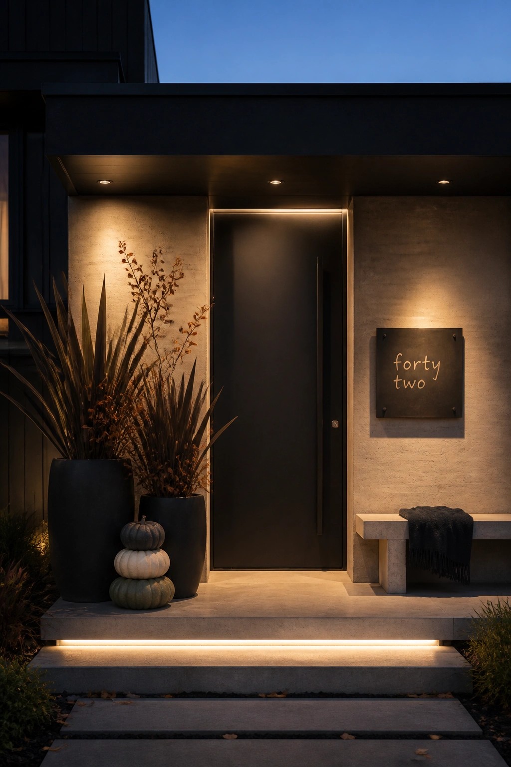 A modern black front door set in a concrete facade with integrated lighting, two large black planters holding dried foliage and stacked gourds, a concrete bench with a dark throw, and a dark metal address plaque reading 