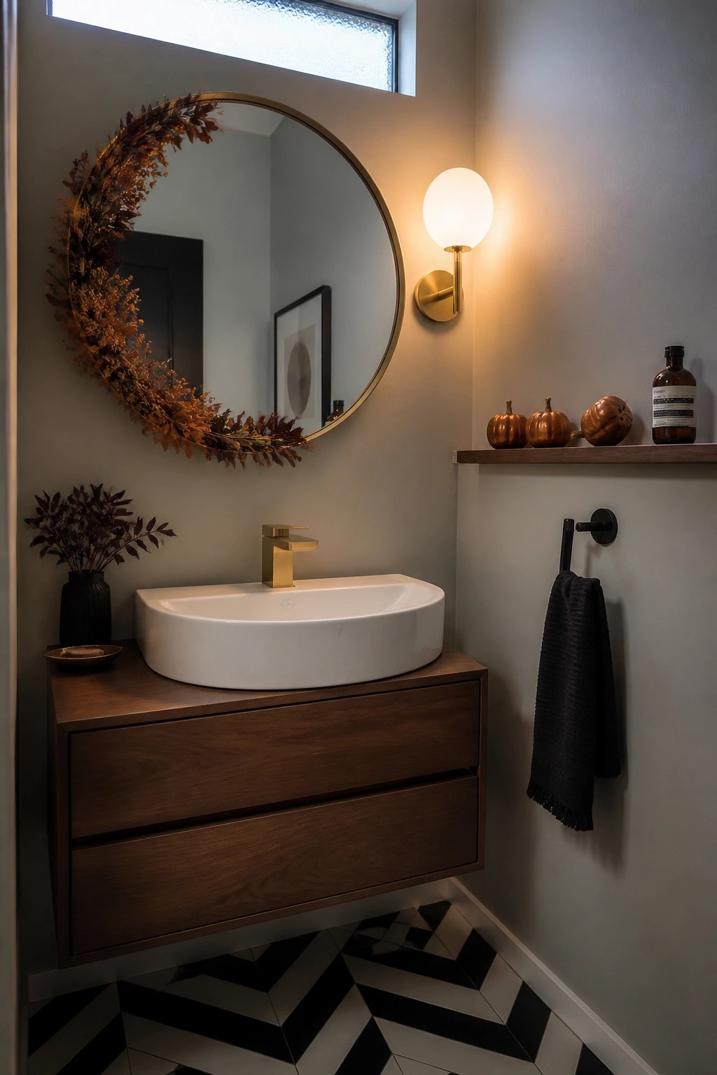 A bathroom with a floating wood vanity, white vessel sink, round mirror with dried leaf wreath, wall sconce, and a narrow shelf holding small pumpkins and a bottle.