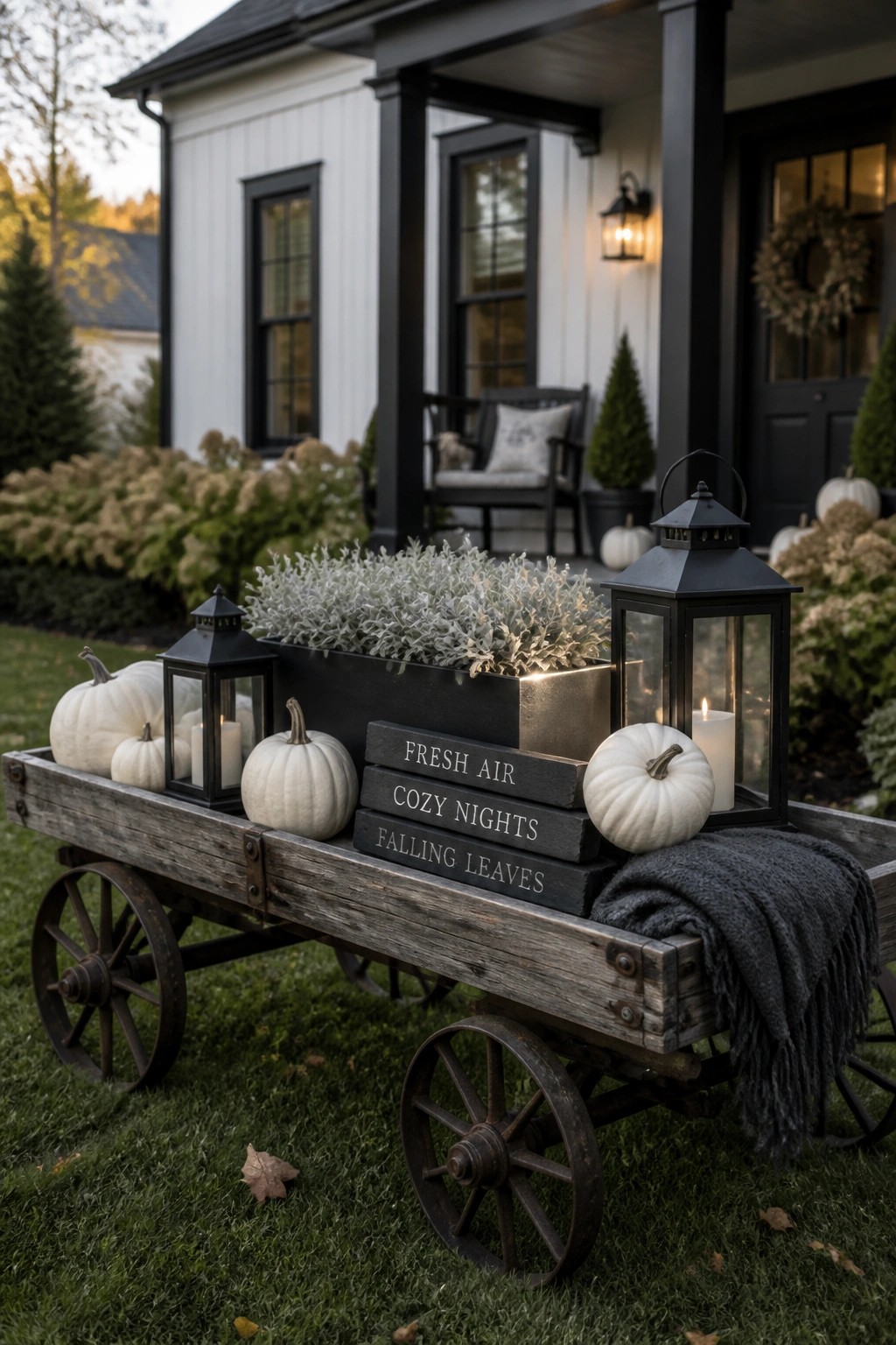 A weathered wooden wagon holding white pumpkins, black lanterns, a metal planter with greenery, and stacked signs sits on the lawn in front of a white house with black trim.
