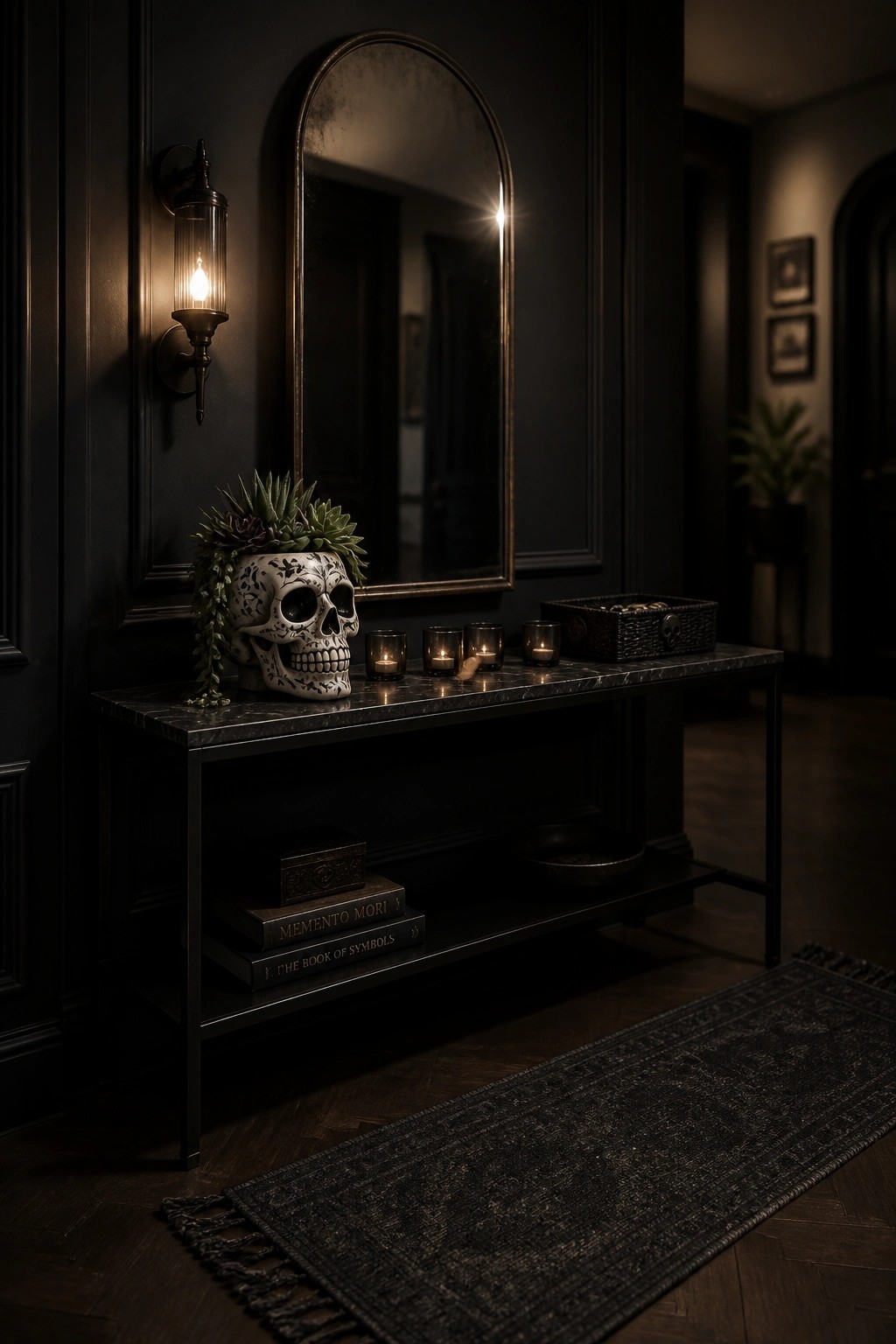 A dark entryway console holds a decorative skull planter filled with succulents, several lit candles, stacked books, and a woven basket beneath an arched mirror on a paneled wall.