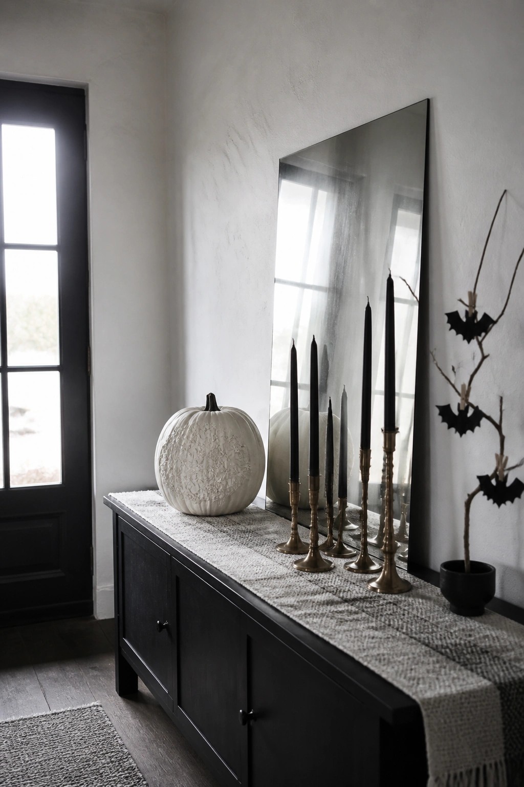 A black console table displays white pumpkins and multiple black taper candles in brass holders, reflected in a large mirror beside a small potted branch with bat decorations.