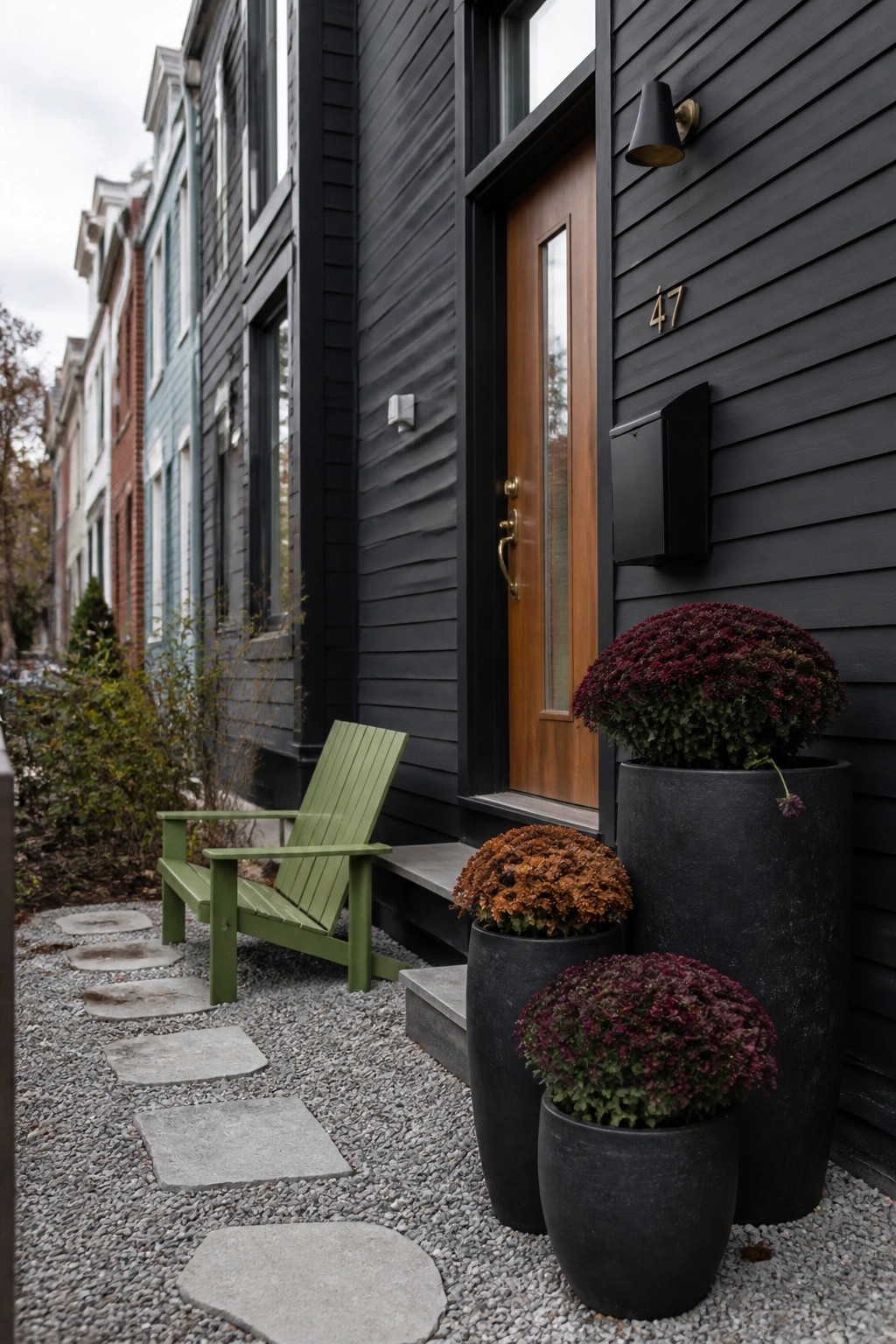 A black house exterior features a wooden front door flanked by three large black pots filled with burgundy and orange chrysanthemums, alongside a green wooden chair on a gravel path with stone stepping stones.