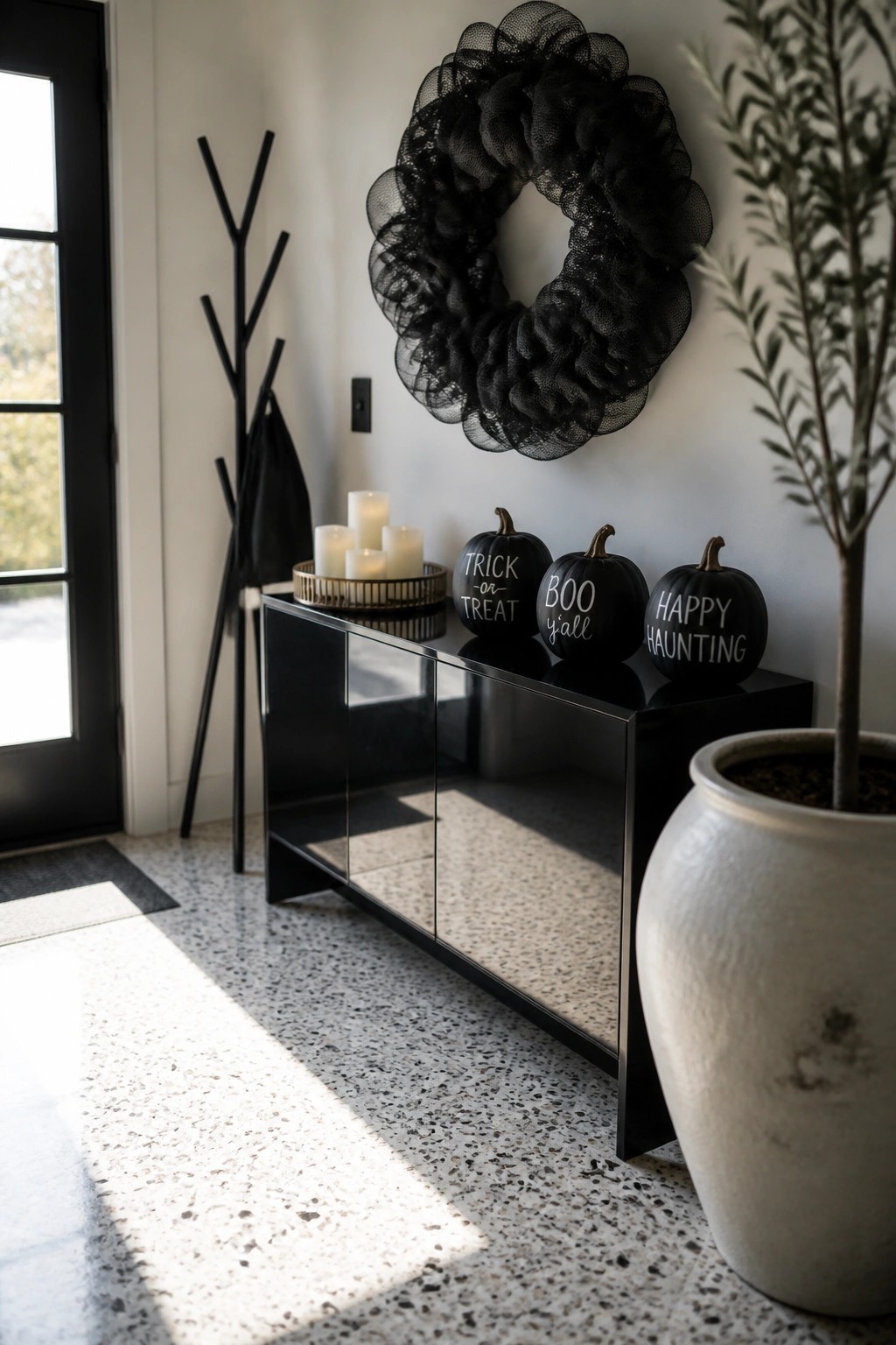 An entryway console holds three black pumpkins with white text, a tray of candles, and a large black mesh wreath hanging on the wall above.