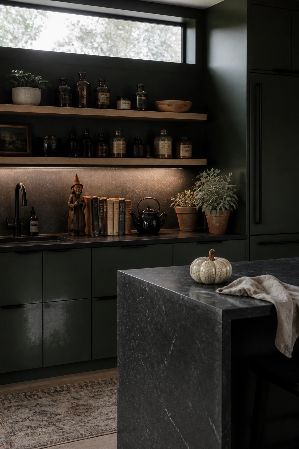 A moody dark green kitchen with open wooden shelves displaying vintage glass bottles, old books, a small witch figurine, and potted plants above a black stone countertop.