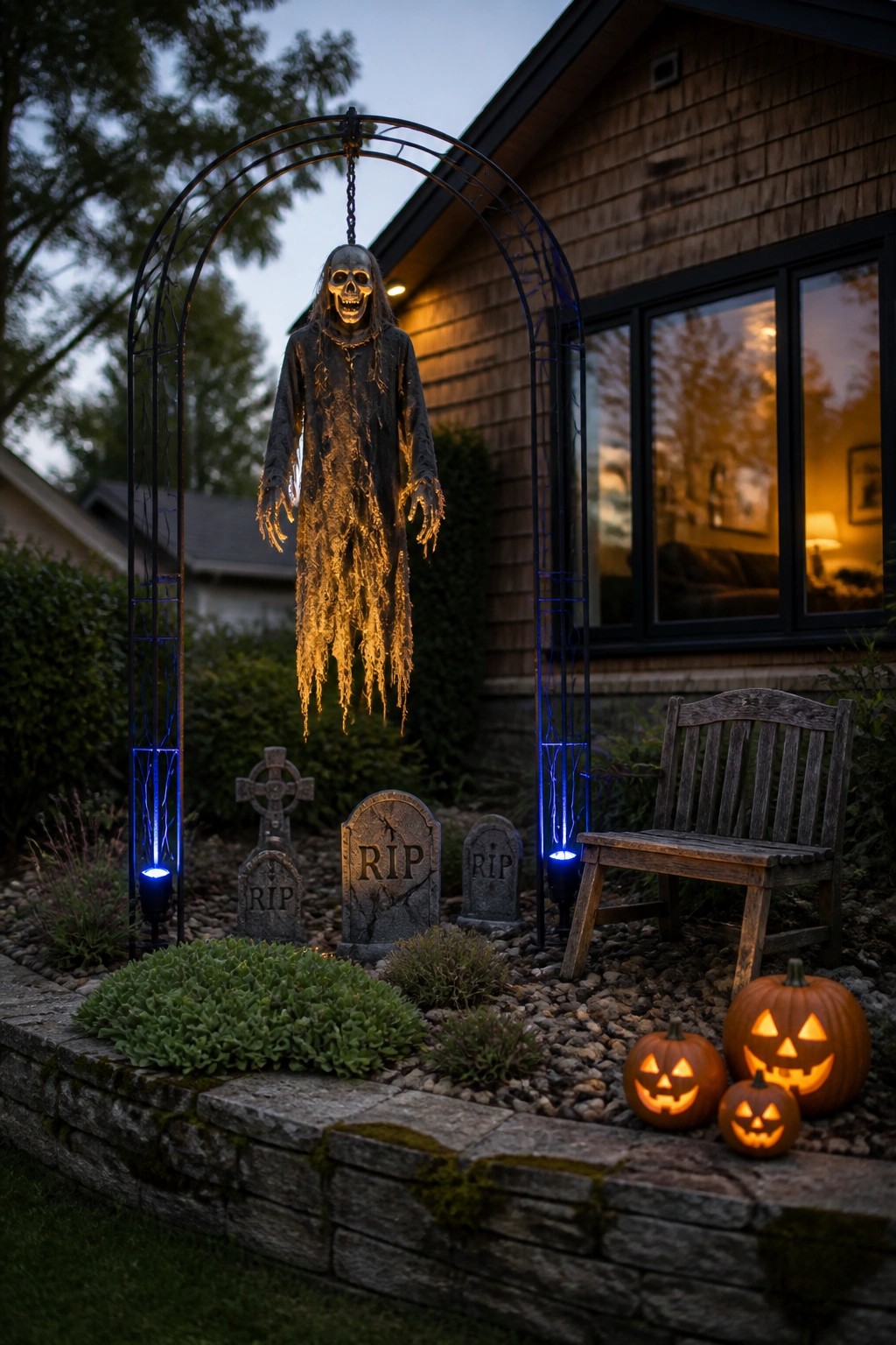 A skeleton figure hangs from a metal garden arch lit with blue lights, with several gravestones and carved pumpkins arranged around the base in a front yard.